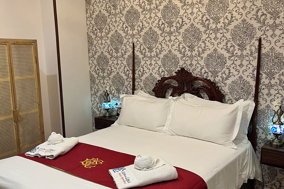 A neatly made bed with white linens, a red throw blanket, and a towel with a logo. There is a wooden headboard and a nightstand with a lamp. The room has patterned wallpaper and a wooden floor.
