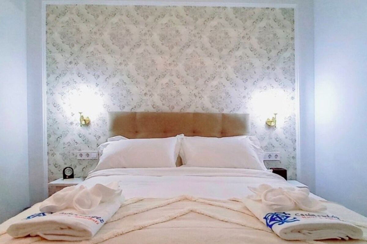 A neatly made bed with a headboard, two bedside lamps, and a folded towel on the side.