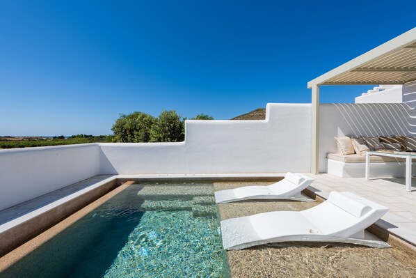 Un área de piscina en la azotea con agua cristalina, sillas de descanso blancas y un edificio de color blanco con un sofá beige.