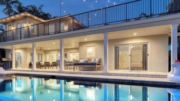 Una casa de dos pisos con piscina, mobiliario exterior y un balcón.