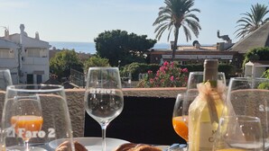 A table set with a bottle of El Faro2 wine, glasses of orange juice, watermelon slices, and a croissant, with a view of the sea and palm trees in the background. - Apartamento Para 4-6 pax en la Costa del Sol, Mijas Málaga. Acepta Mascotas (AN)
