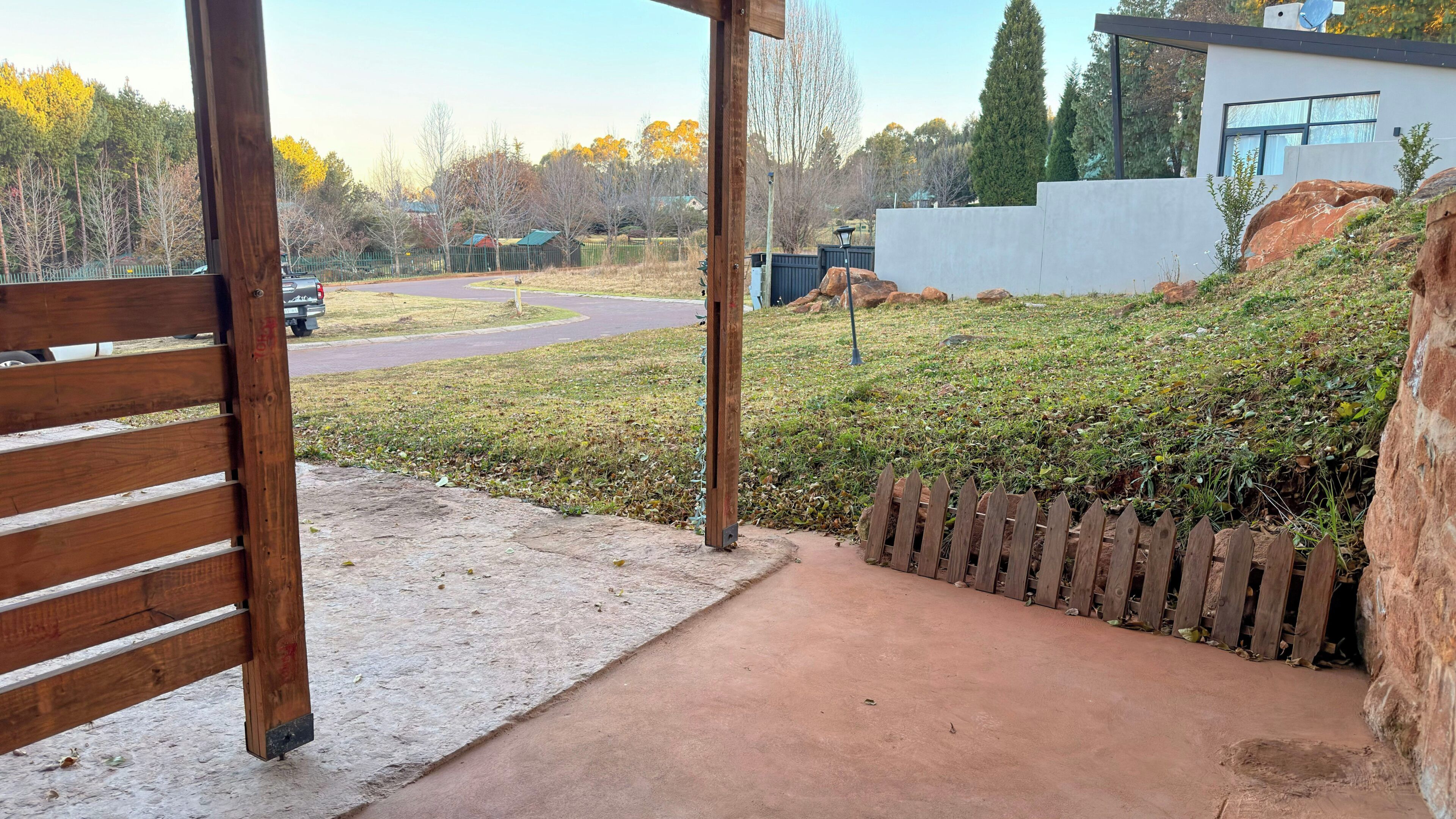 Een overdekt buitengebied met een betonpad dat naar een modern gebouw leidt, omgeven door een houten omheining en een grasheuvel.