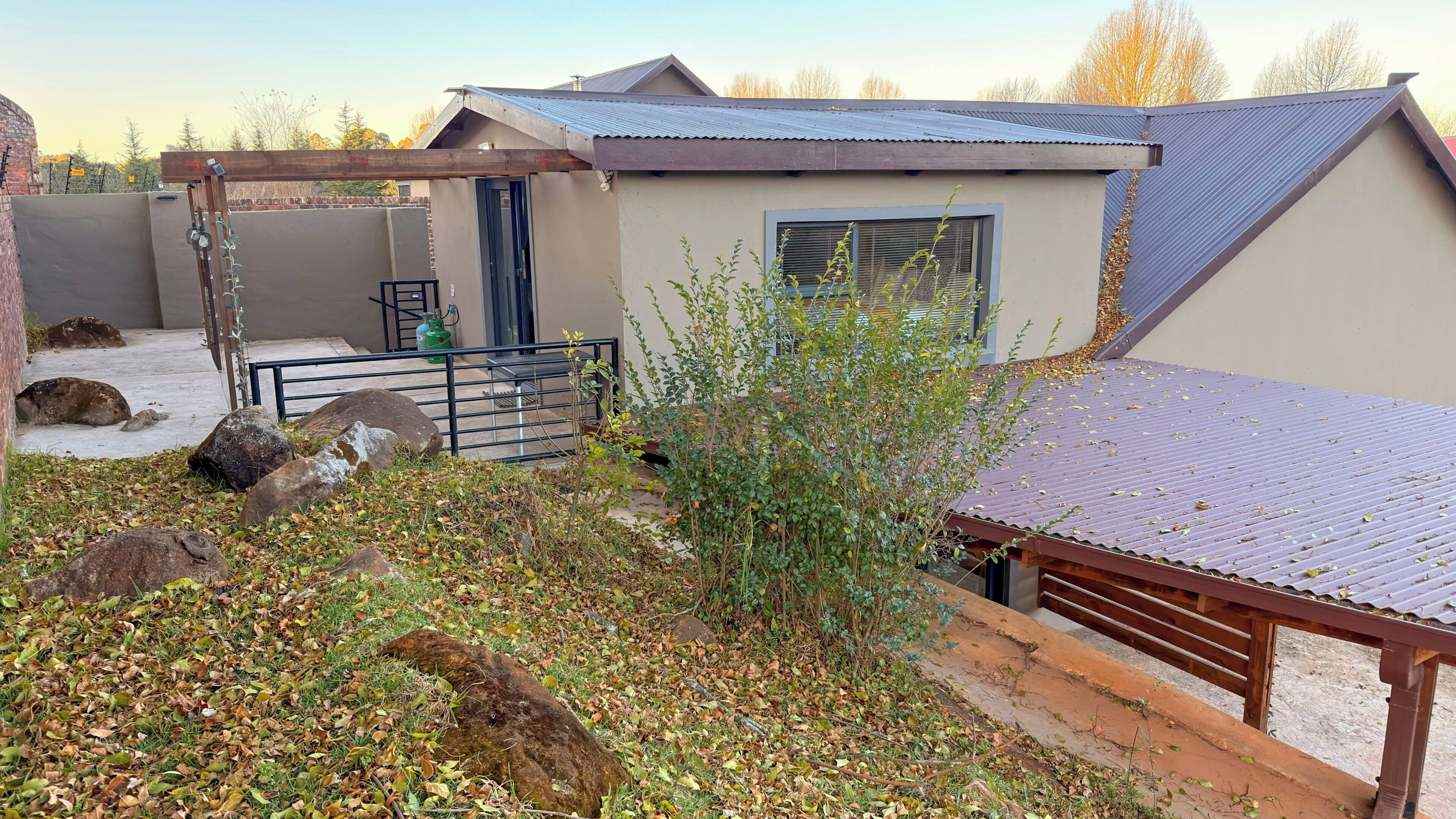 Una vivienda con techo de metal, un pequeño jardín con rocas y plantas, y un cilindro de gas a la vista.