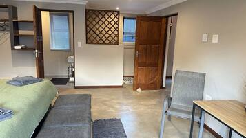 A room with a bed, a desk, a chair, and a small table. There is a window with blinds, a door, and a shelf with items on it.
