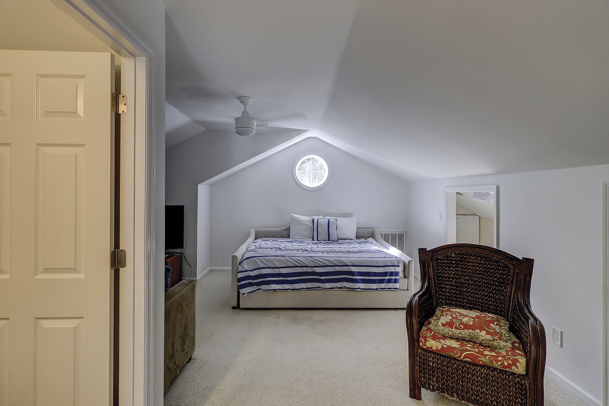 A bedroom with a sloped ceiling, a bed with striped bedding, a wooden chair with a cushion, and a small window.