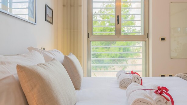A neatly made bed with white linens and pillows, a window with blinds, and a towel with a red ribbon on the bed.