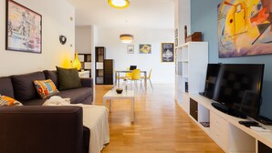 A modern living room with a sofa, coffee table, and TV. There is a dining area with chairs and a table in the background.