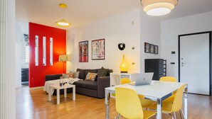A modern living room with a red accent wall, a sofa, a coffee table, and a dining area with yellow chairs and a laptop.