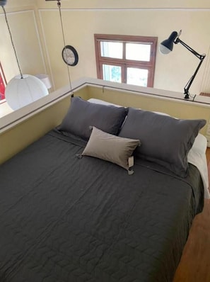 Uma cama com um edredom escuro e duas almofadas, uma janela com vista e uma luminária de mesa.