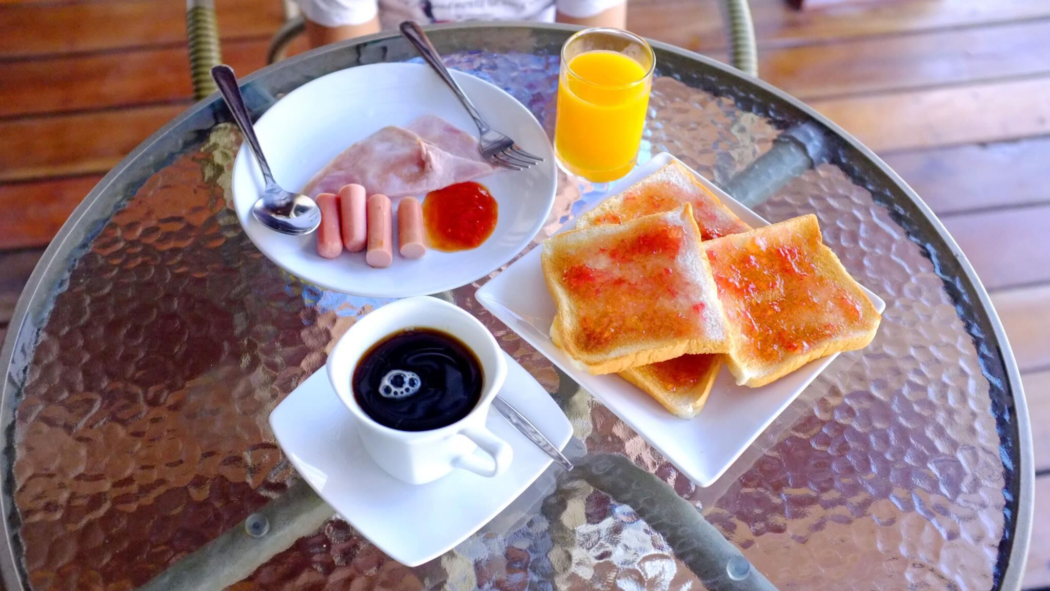 Mesa de vidro com torrada, geleia, linguiça, copo de suco de laranja e uma xícara de café.
