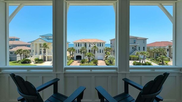 A room with a view of a large building with a red roof and palm trees. There are three Adirondack chairs in the foreground. - Sea Pearl- Jewel of Gulf Shores! Community pool (Gulf Shores)
