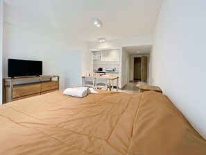 Un dormitorio con una cama grande, un televisor sobre un mueble de madera, una kitchenette y un baño visible a través de una puerta abierta.