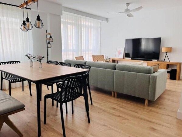 Una sala de estar moderna con una mesa de comedor de madera, sillas negras, un sofá gris y un televisor de pantalla plana.
