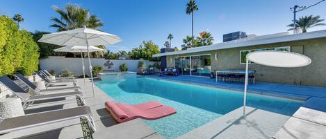 A modern outdoor pool area with lounge chairs and umbrellas, surrounded by palm trees and a clear blue sky.