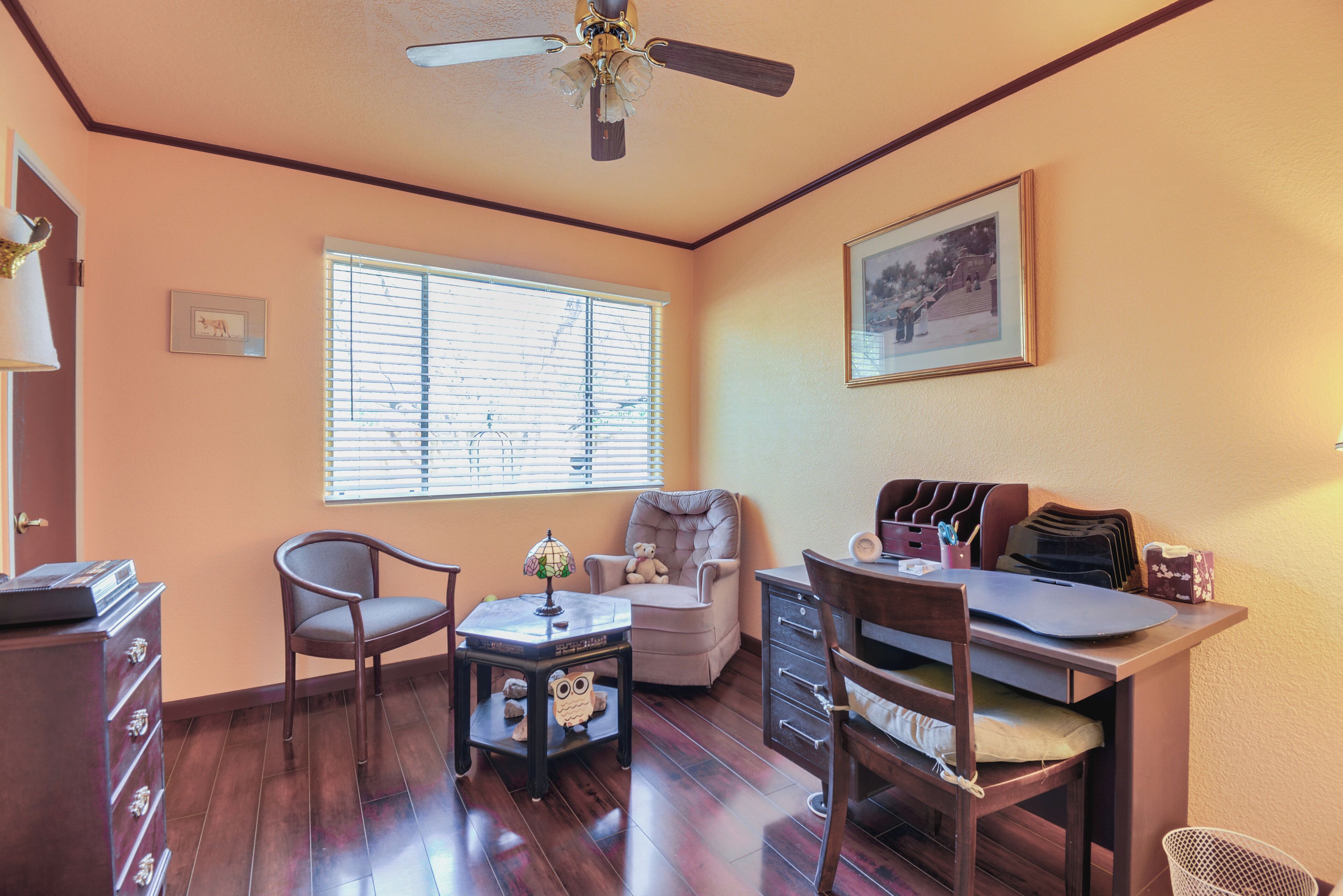 A room with a ceiling fan, a window with blinds, a desk with a chair, a dresser, and a framed picture on the wall.