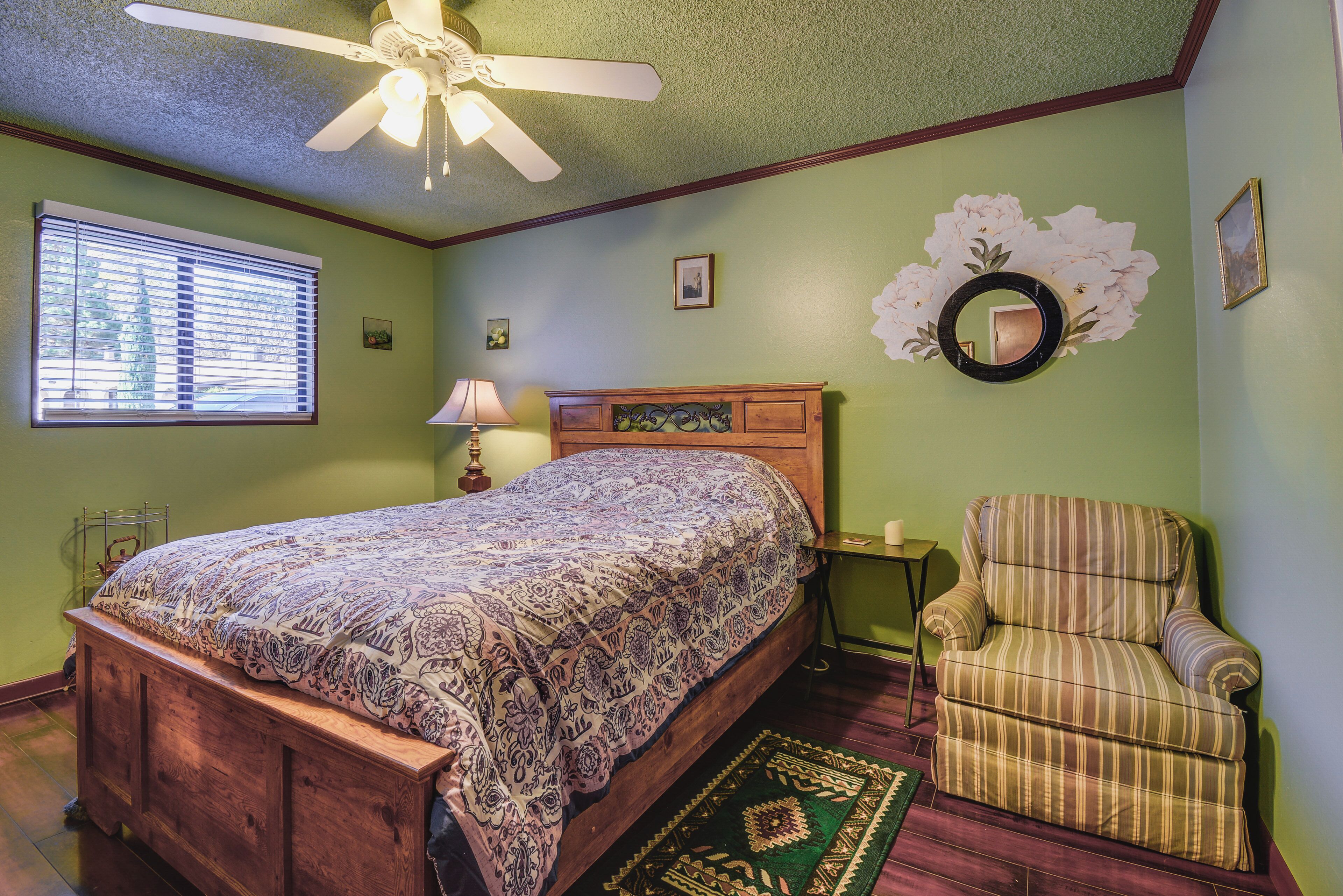 A bedroom with a wooden bed, a green wall, a ceiling fan, a window with blinds, a chair, and a mirror.