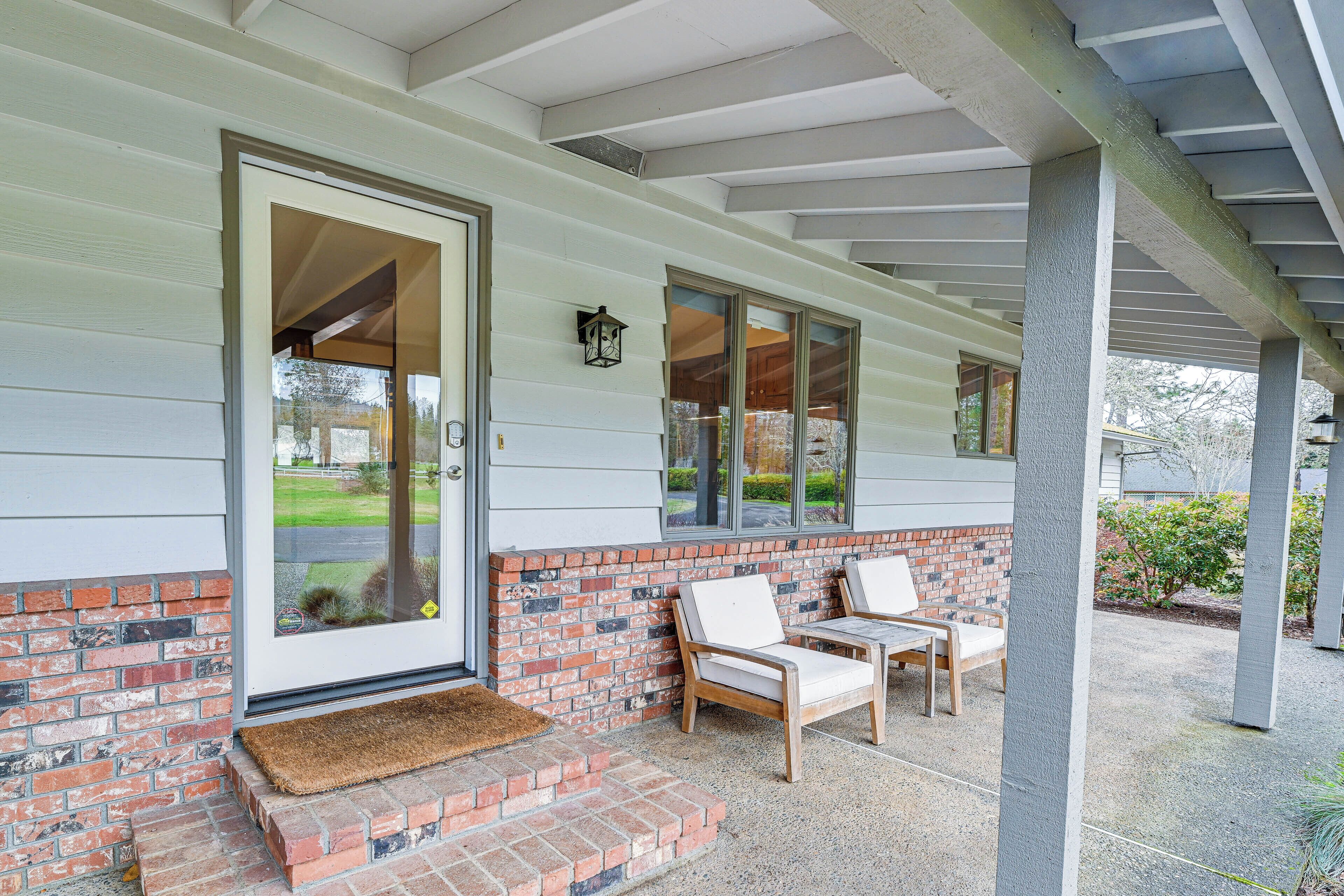 A covered porch with two white cushioned chairs, a brick wall, and a door with a doormat.