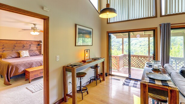 A bedroom with a bed, a desk with two stools, and a balcony with a view. - Maggie Valley Mountain Escape w/ Fireplace & Deck! (Maggie Valley)