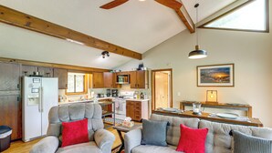 A spacious living area with a grey sofa set, wooden beams, a kitchen with modern appliances, and a large window providing natural light. - Maggie Valley Mountain Escape w/ Fireplace & Deck! (Maggie Valley)