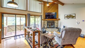 A spacious living room with a fireplace, a large TV, and a balcony with a view of the outdoors. - Maggie Valley Mountain Escape w/ Fireplace & Deck! (Maggie Valley)