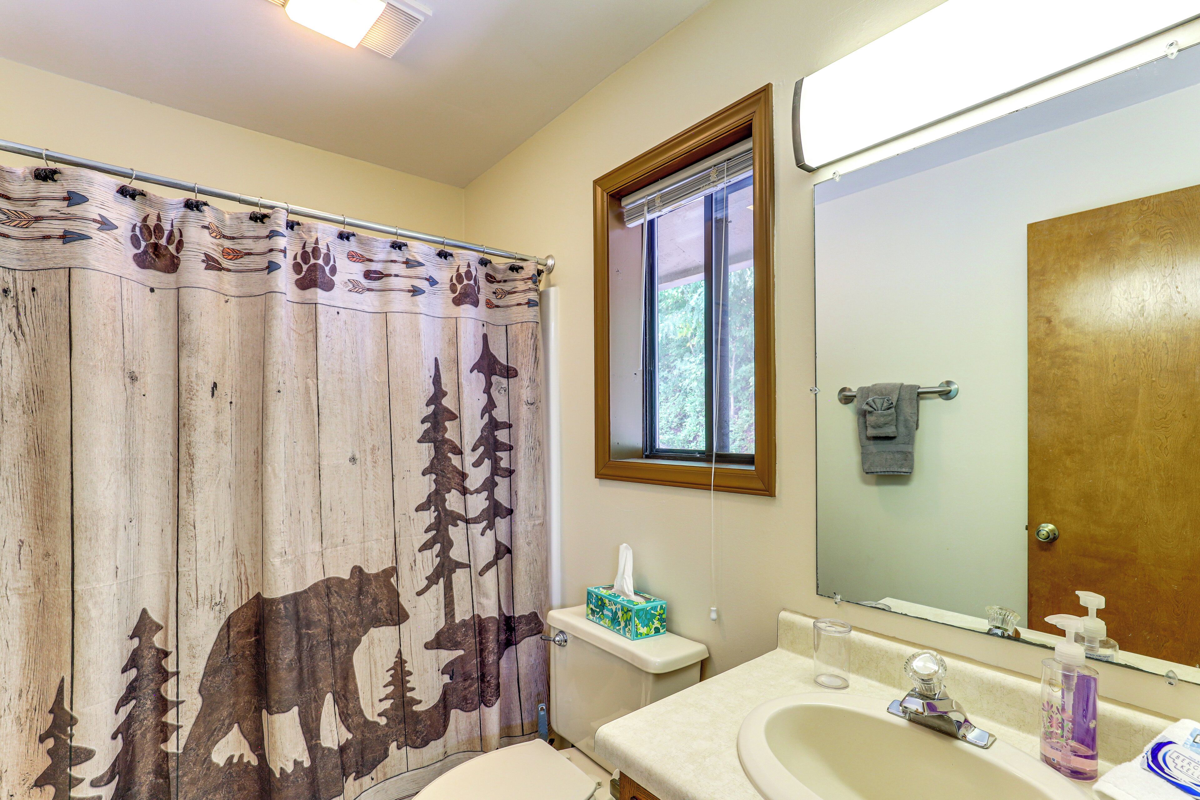 Une salle de bain avec un rideau de douche en bois orné de motifs animaliers, des toilettes, un lavabo avec un miroir et une fenêtre avec des stores.
