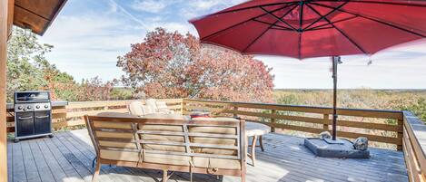 A wooden deck with a bench and a grill, a red umbrella, and a view of trees and landscape.