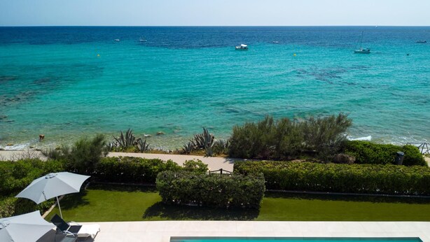 A view of a pool area with a clear blue sea in the background, surrounded by greenery and a few boats.