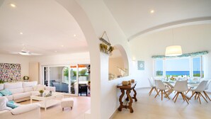 A spacious living area with a white sofa, coffee table, and ottoman. There is a dining area with a table and chairs, and a view of the ocean through large windows.