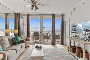 A spacious living room with a large television, a sofa, and a coffee table. The room has a view of the beach through large glass doors. - Blue Sky-Gulf front resort w/pool, gorgeous views! (Navarre)