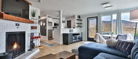A modern living room with a fireplace, a sofa, and a view of the mountains through large windows.