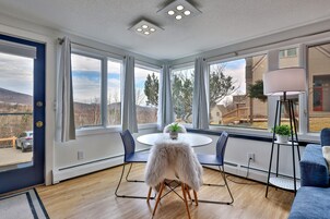 Una habitación con ventanas amplias, una mesa redonda con sillas, un sillón blanco y mullido, y una lámpara de pie.