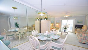 A spacious living area with a glass dining table surrounded by white wicker chairs, a sofa with floral upholstery, and a television in the background. - Baywatch Iii 302a 2 Bedroom Condo by RedAwning (Ocean City)