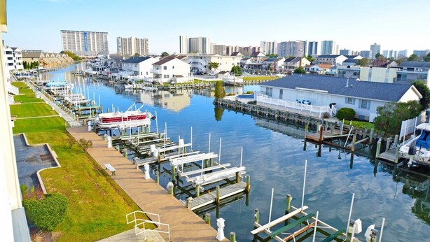 A marina with docked boats, a waterfront property with a house, and high-rise buildings in the background. - Baywatch Iii 302a 2 Bedroom Condo by RedAwning (Ocean City)