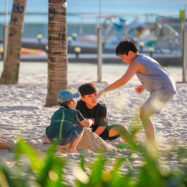 Tres personas jugando con arena en una playa con palmeras y un cielo despejado al fondo.