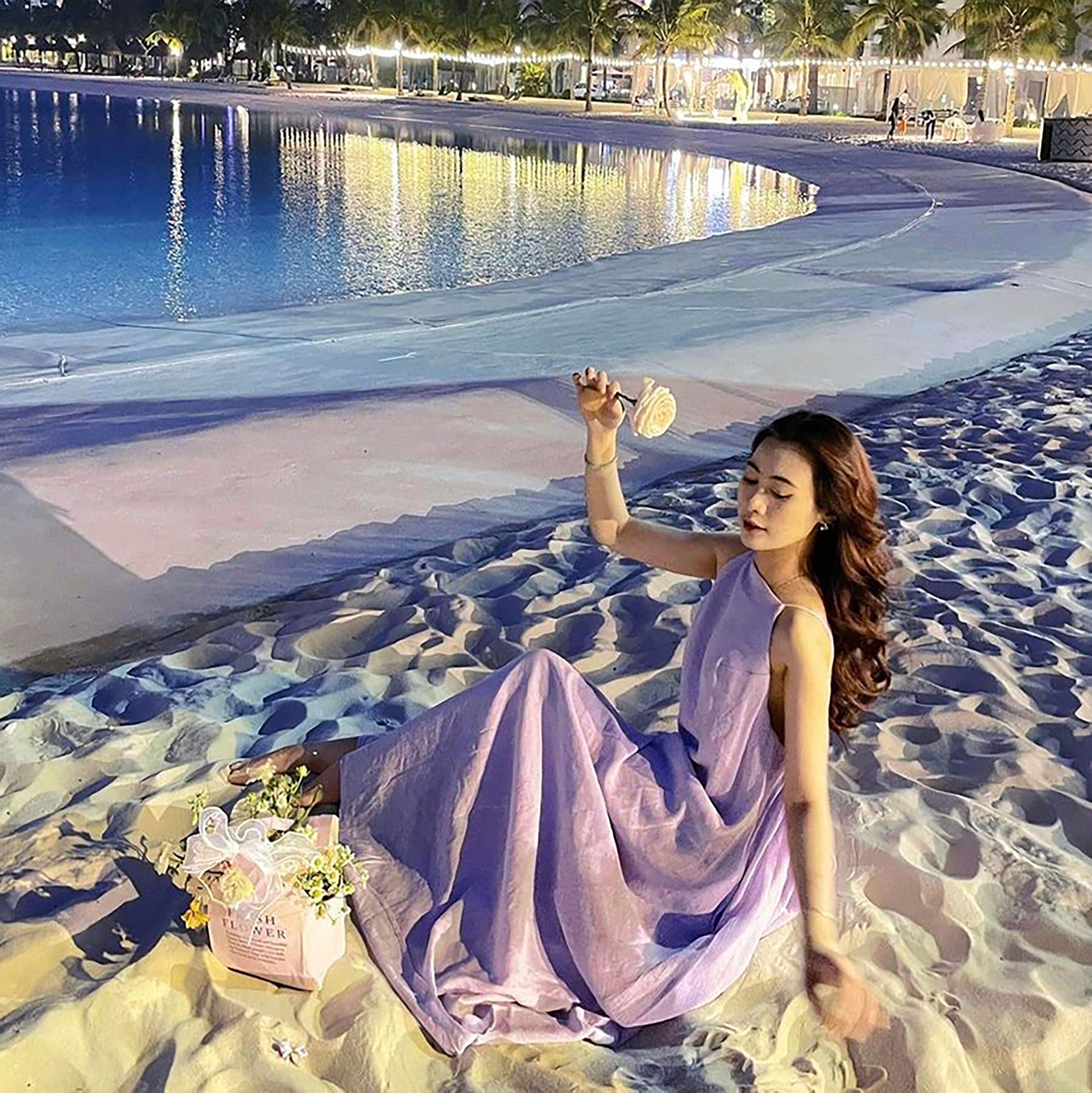 A person sitting on a sandy beach near a pool with a bag of flowers.