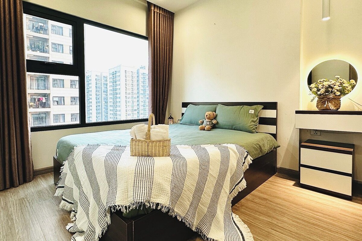 A bedroom with a bed, a bedside table, a dresser, a window with curtains, and a view of buildings outside.