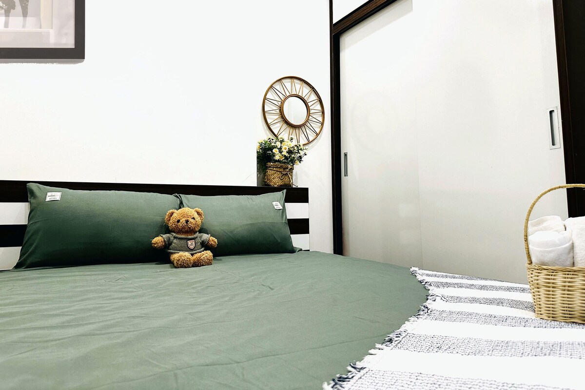 A neatly made bed with a green bedspread and a teddy bear on the pillow. There is a wall clock and a small plant on the wall.