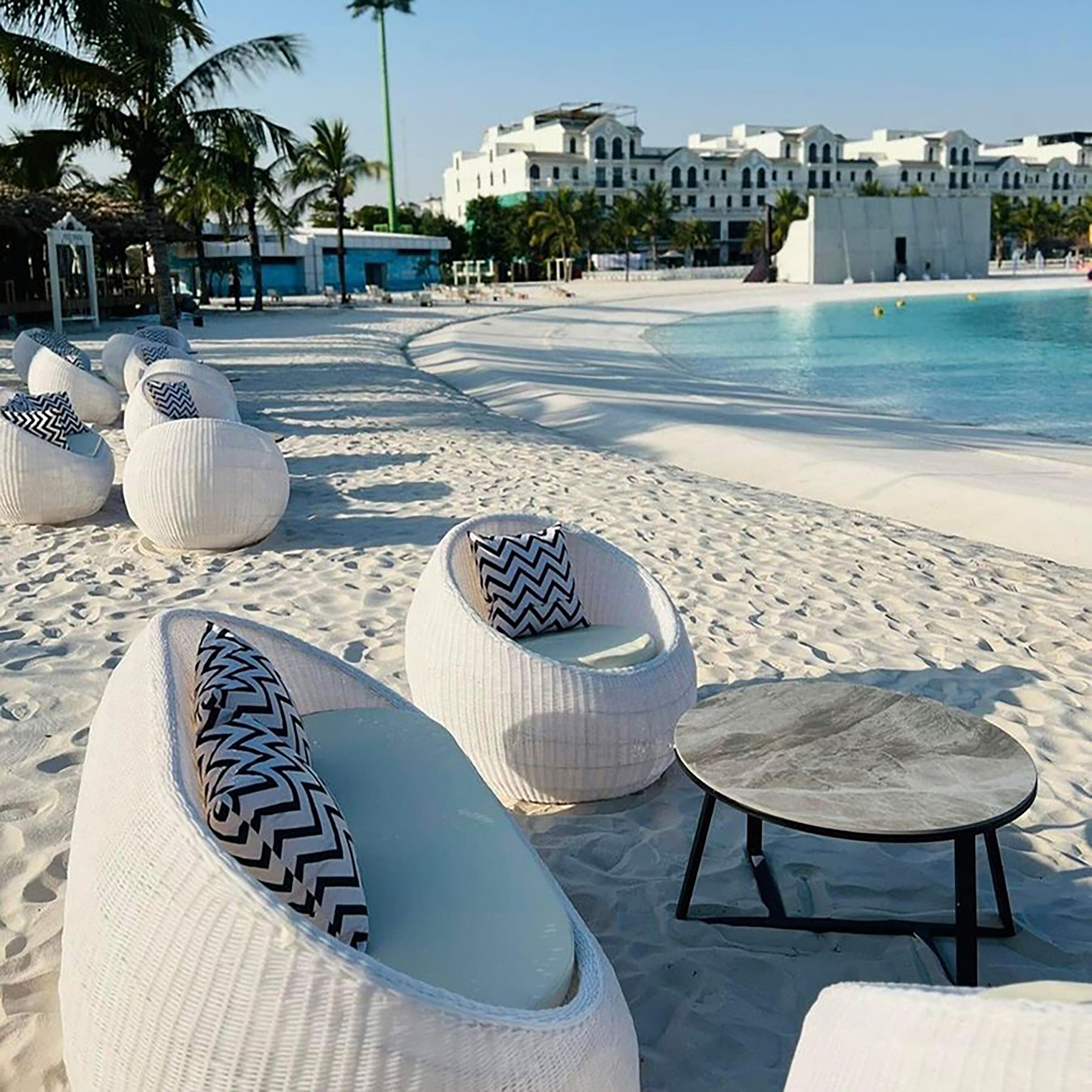 A beachside lounge area with white cushioned seating and a round table, overlooking a pool and a building with multiple stories.
