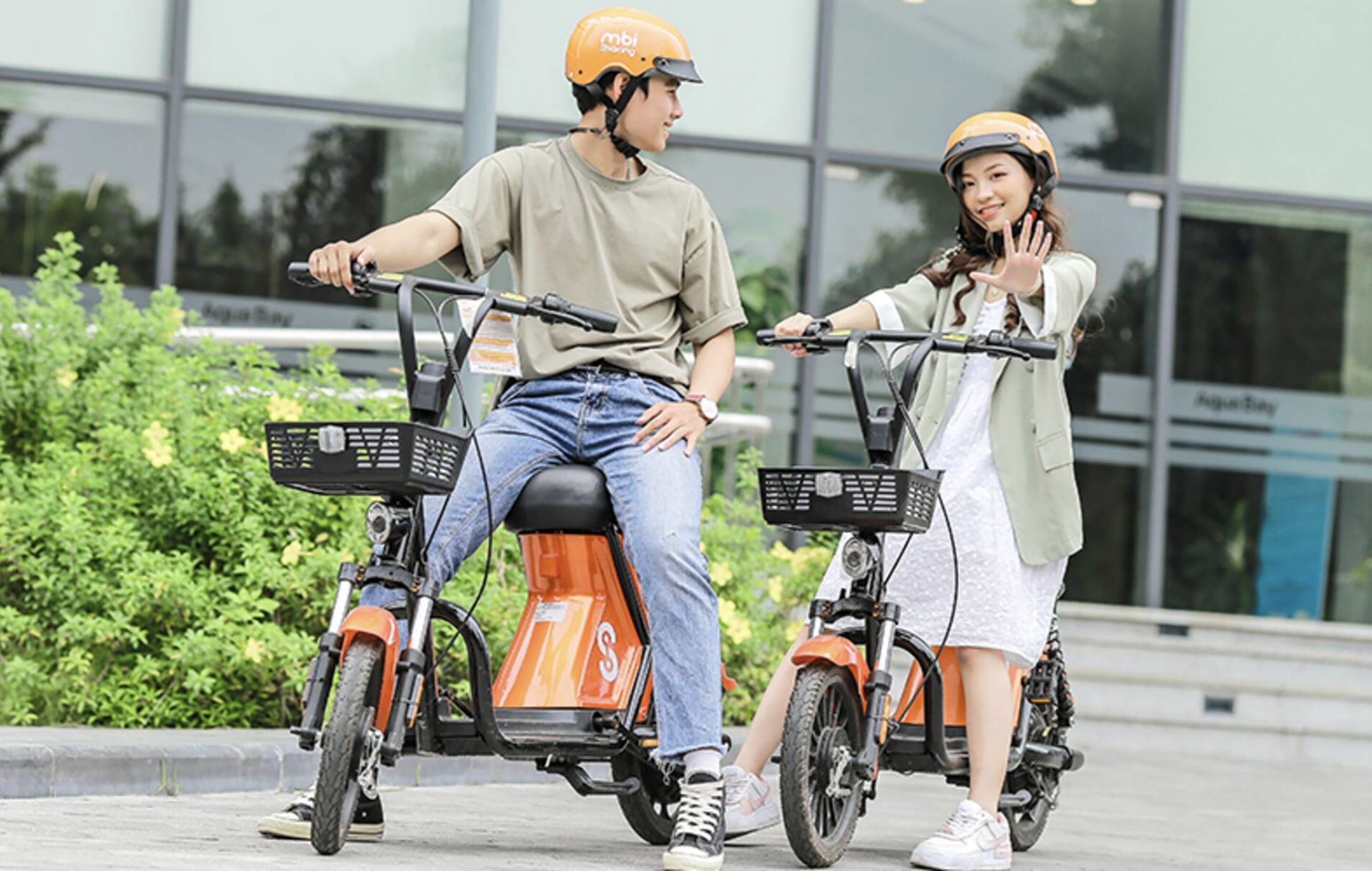 Two people on electric scooters with baskets, in front of a building with glass windows.