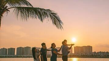 A sunset view with a group of people on a beach, palm trees, and a city skyline in the background.