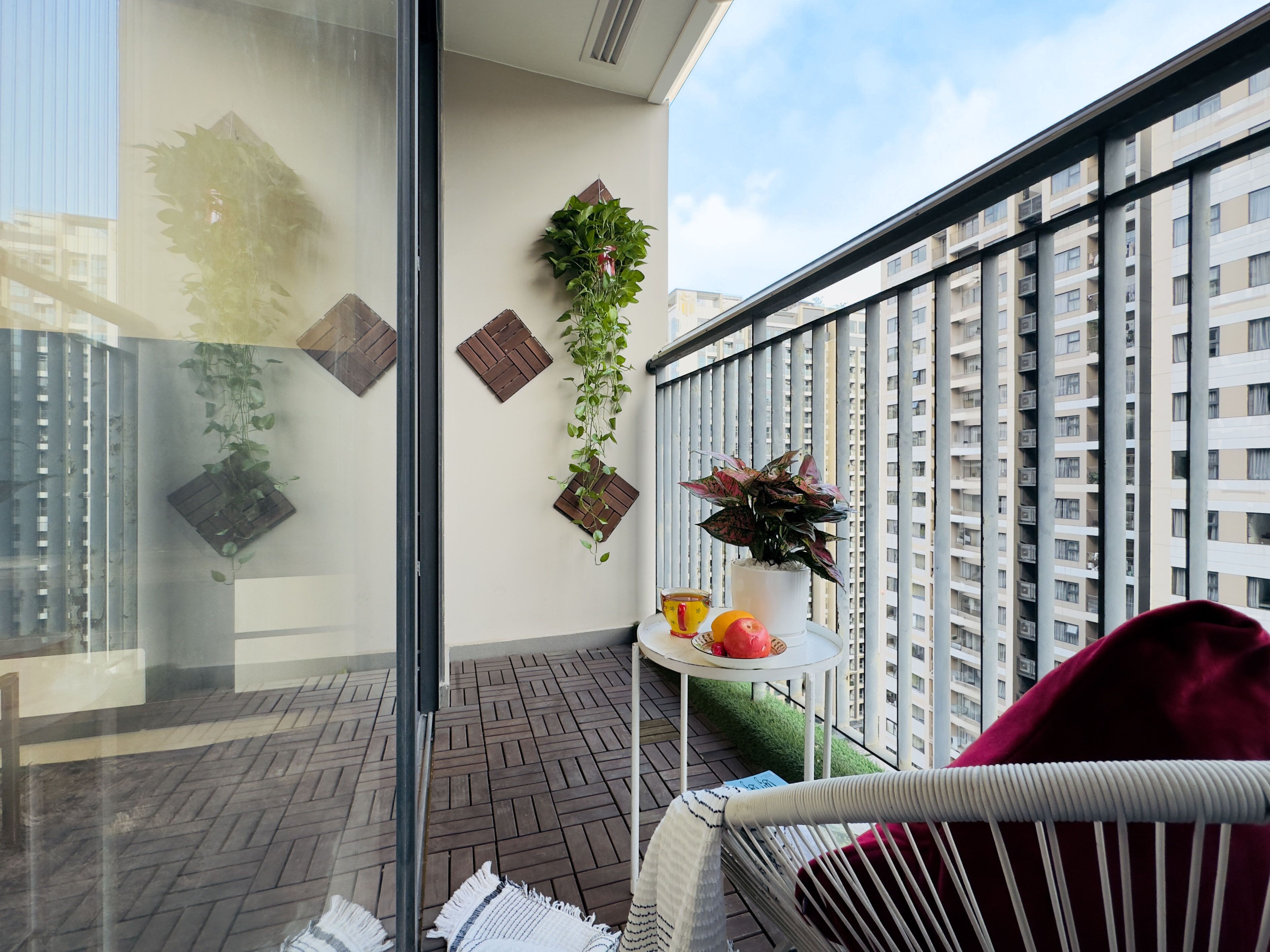 Un balcon donnant sur un immeuble de grande hauteur, avec des plantes en pot, une petite table sur laquelle se trouvent des fruits et une tasse, et une chaise en osier.