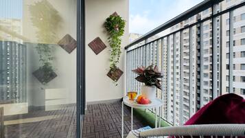 Un balcon donnant sur un immeuble de grande hauteur, avec des plantes en pot, une petite table sur laquelle se trouvent des fruits et une tasse, et une chaise en osier.
