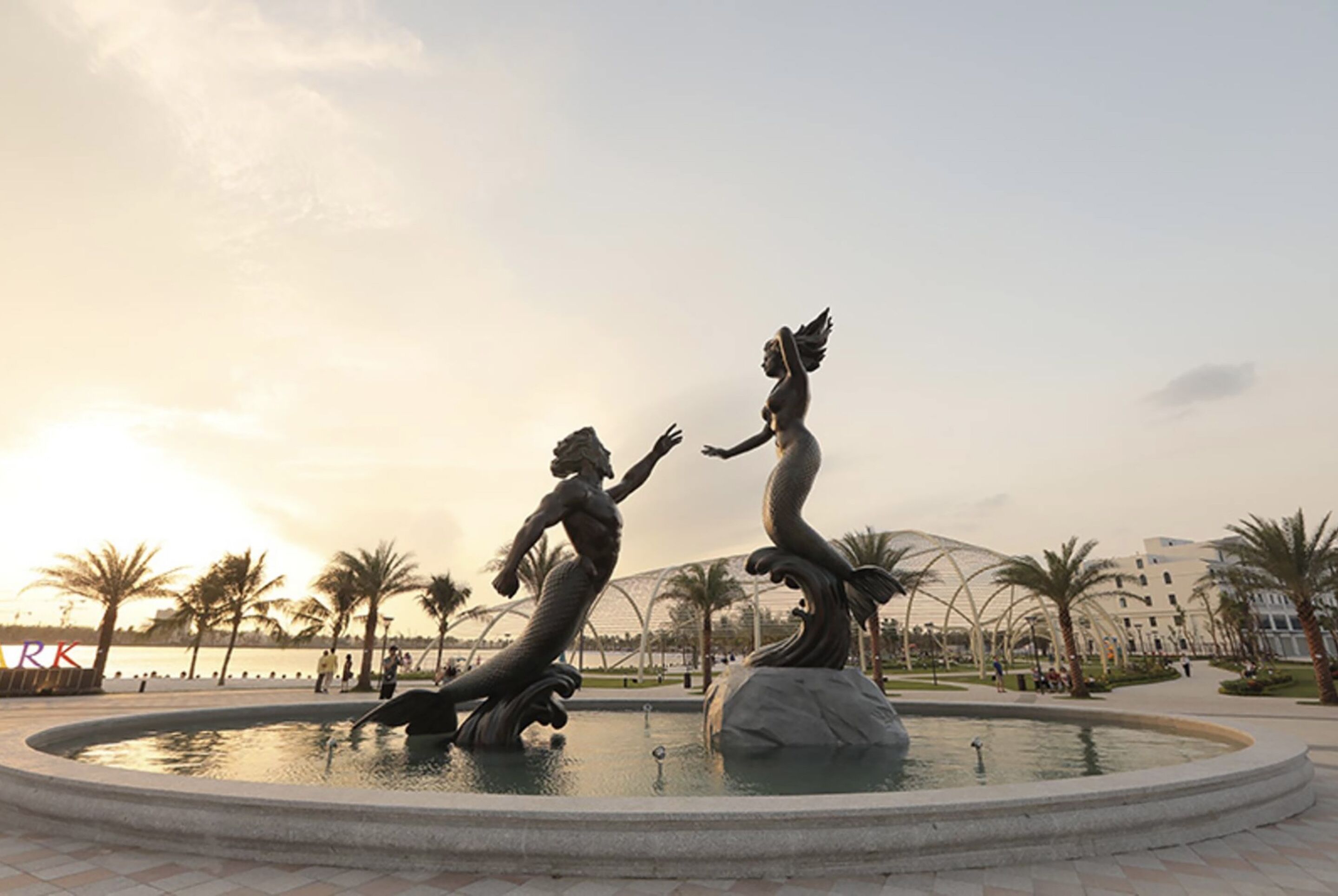 A fountain with two statues of mermaids in a circular pool, surrounded by palm trees and buildings in the background.