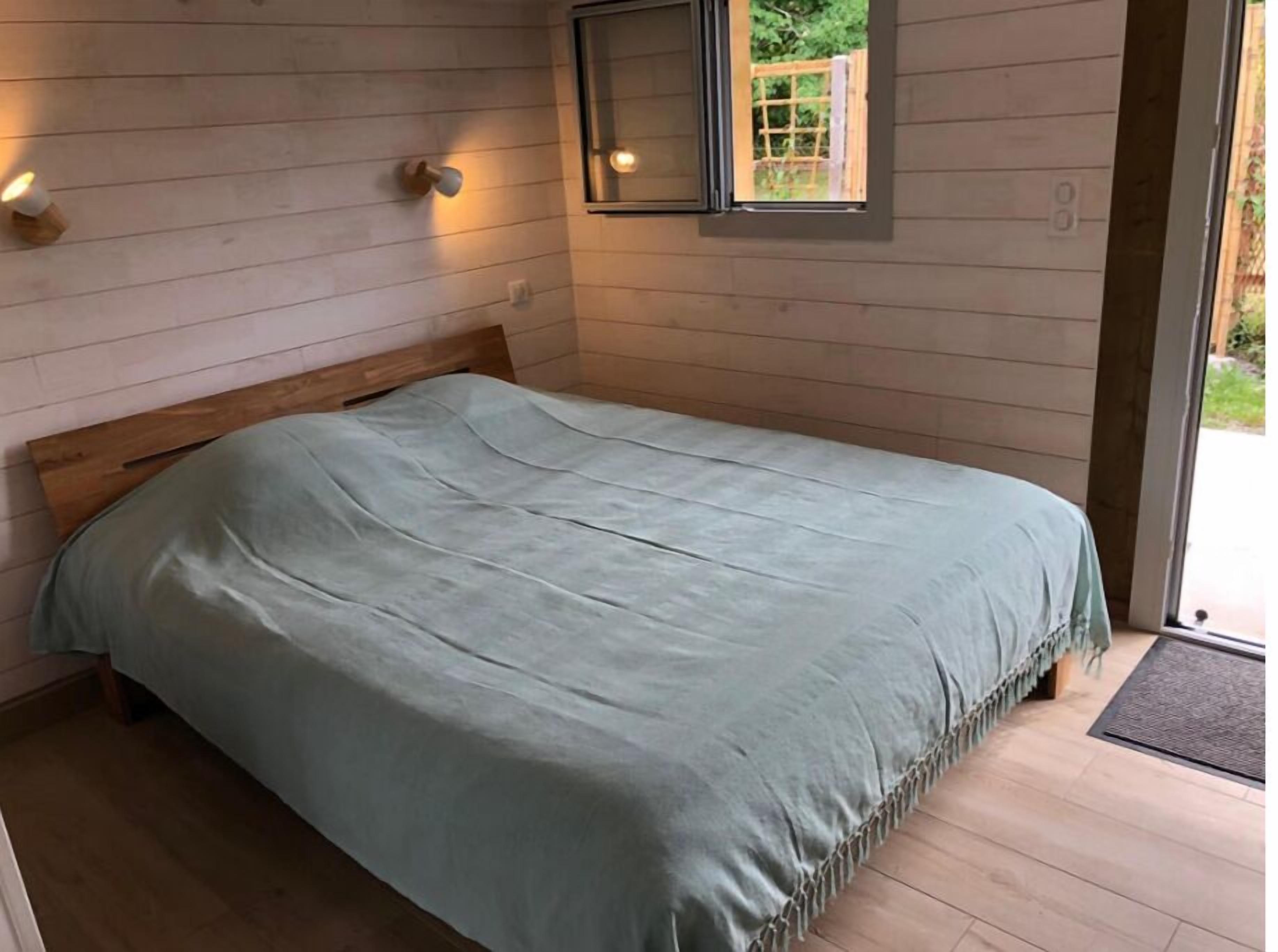 A bedroom with a wooden headboard, a light blue bedspread, and a small window with a view of greenery outside.