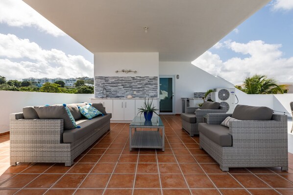 Un patio cubierto al aire libre con mobiliario de mimbre, piso de baldosas y una zona de cocina con salpicadero de piedra.
