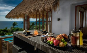 Une table à manger avec des aliments pour le petit-déjeuner, un panier de fruits et une vue sur l’océan.