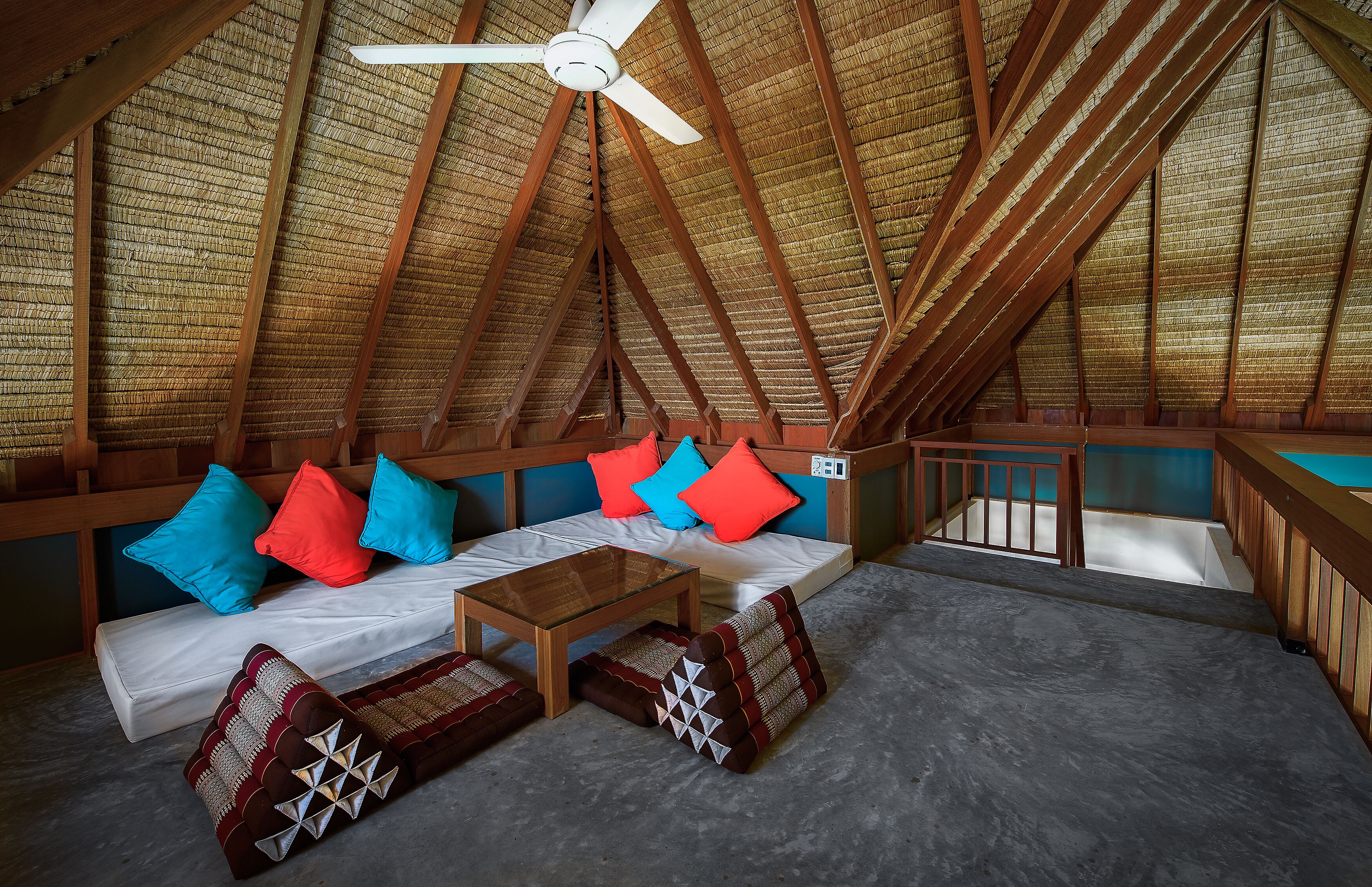 Une chambre mansardée avec un plafond en pente, des poutres en bois, un ventilateur de plafond, un canapé avec des coussins, une petite table et un escalier.