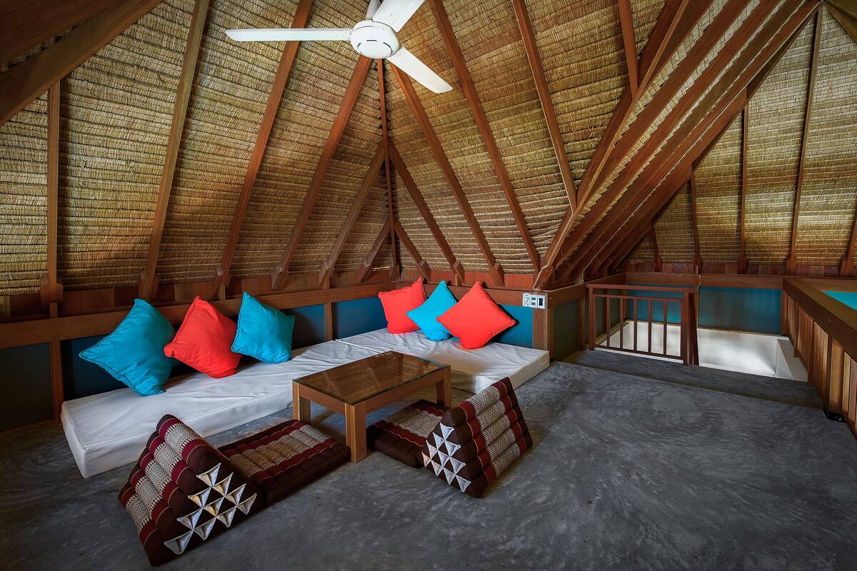 An attic room with a sloped ceiling, wooden beams, a ceiling fan, a sofa with cushions, a small table, and a staircase.