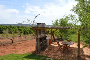 An outdoor barbecue area with a grill, dining table, and chairs, surrounded by trees and a vineyard in the background.