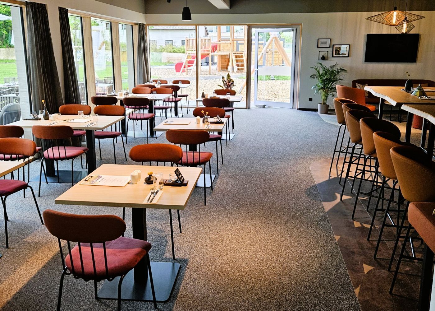 A spacious indoor dining area with multiple tables and chairs, large windows, and a view of an outdoor play area.